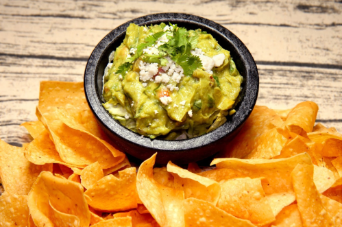 A bowl of guacamole topped with cheese and cilantro, surrounded by crispy tortilla chips on a wooden surface.