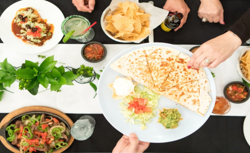 A table spread with various Mexican dishes, including quesadillas, chips, salsa, and drinks, with hands reaching for food.
