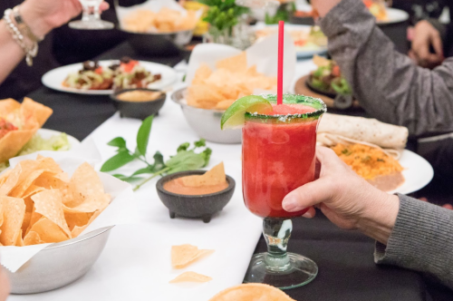 A hand holds a vibrant red cocktail with a lime slice, surrounded by chips and various dishes on a dining table.