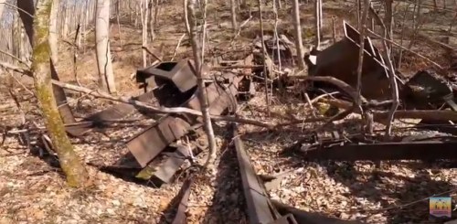 Abandoned metal structures and debris scattered among trees and leaves in a wooded area.