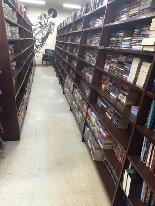 A narrow aisle between tall bookshelves filled with various books in a library, with a chair at the end.