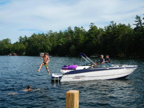 A boy jumps off a boat into a lake while two people sit on the boat and another swims nearby, surrounded by trees.