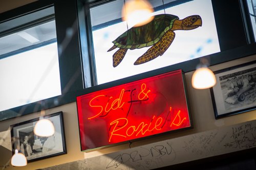 Neon sign reading "Sid & Roxie's" with a turtle decoration, set in a cozy restaurant interior.