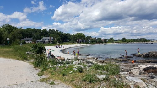 A sandy beach with people enjoying the water, surrounded by greenery and houses under a partly cloudy sky.