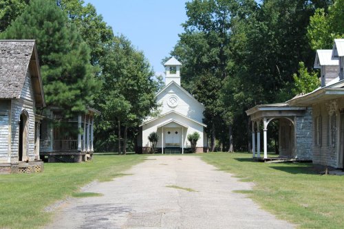 A gravel path leads to a white church surrounded by trees and old wooden buildings on either side.