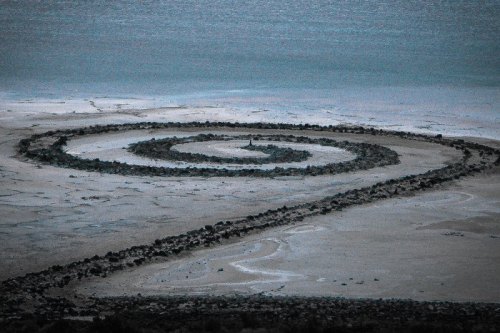 A spiral stone formation on a sandy landscape near a body of water, with a calm, muted color palette.