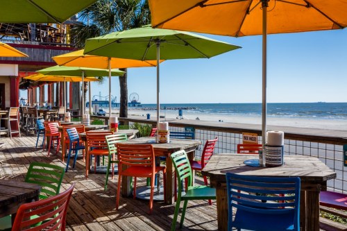 Colorful outdoor seating with umbrellas overlooking a beach and ocean, with a boardwalk and amusement rides in the distance.