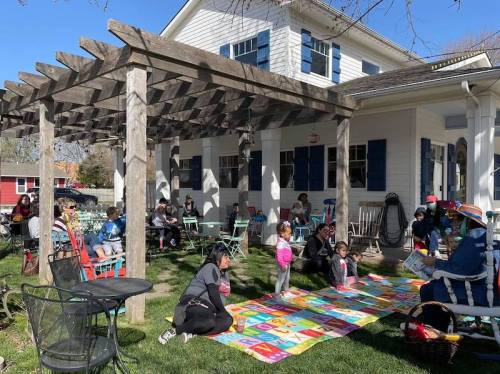 A group of children and adults gather outdoors under a pergola, enjoying a storytime on a colorful blanket.