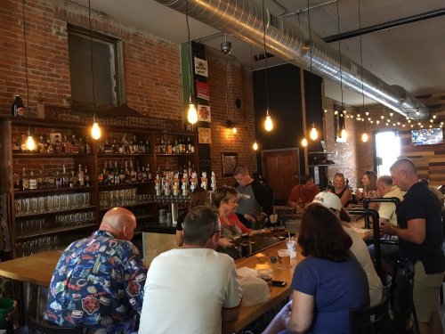 A lively bar scene with patrons at the counter, exposed brick walls, and warm lighting creating a cozy atmosphere.