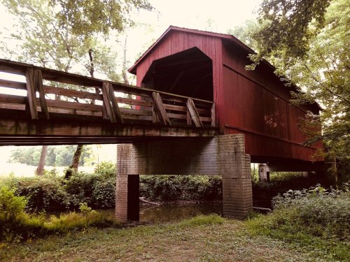 A red covered bridge spans over a small stream, surrounded by lush greenery and trees.