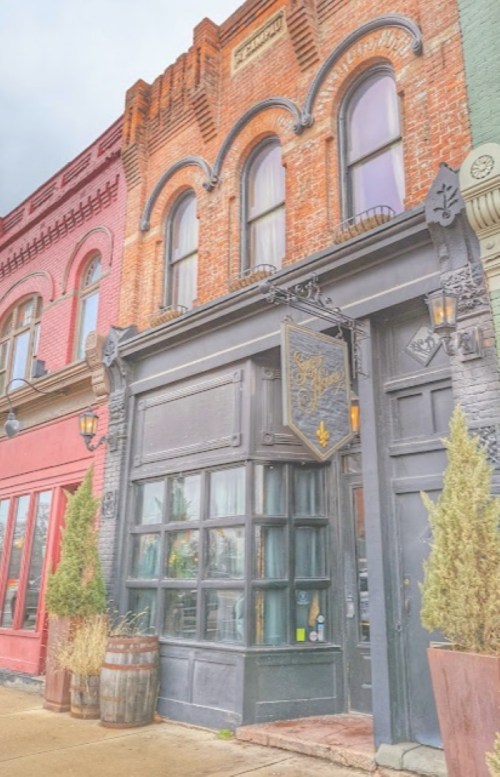 Historic brick building with large windows, a black door, and decorative signage, flanked by potted plants.