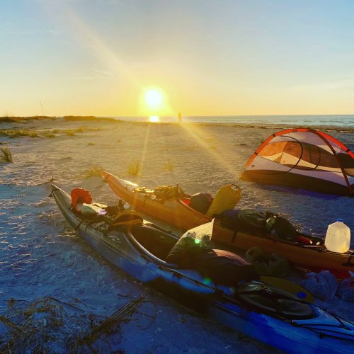 Kayaks on a sandy beach at sunset, with a tent nearby and a person walking along the shore in the distance.