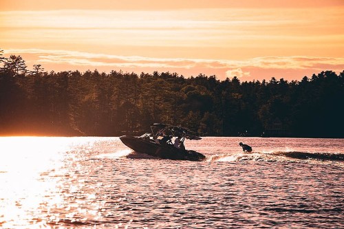 A boat pulls a wakeboarder across a shimmering lake at sunset, surrounded by trees and a colorful sky.