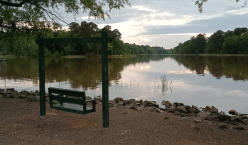 A green swing by a calm lake at sunset, surrounded by trees and rocky shoreline.