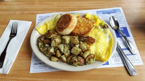 A plate of breakfast featuring an omelet, roasted potatoes, a toasted English muffin, and a side of fruit.