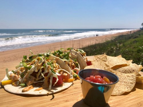 Tacos with toppings and salsa on a wooden table, with a beach and ocean in the background under a clear blue sky.