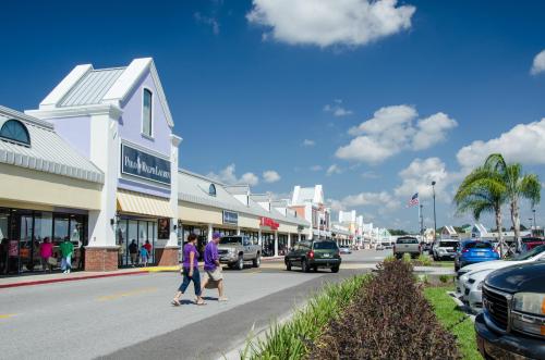 A shopping plaza with people walking, cars parked, and palm trees under a blue sky with fluffy clouds.