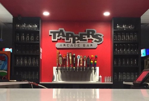 A bar with a red wall featuring the "Tappers Arcade Bar" sign and a row of colorful beer taps.