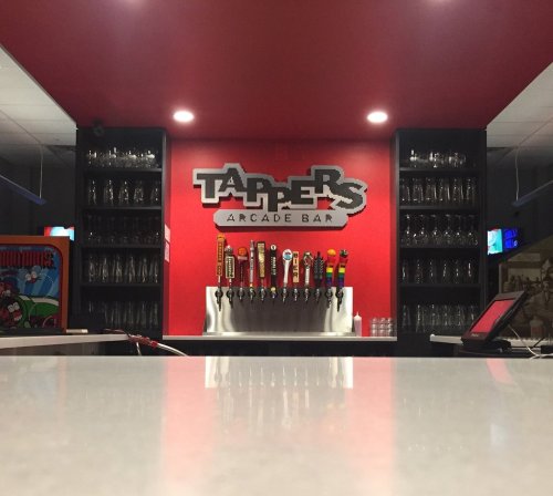 A bar with a red wall featuring a sign that reads "Tappers Arcade Bar" and multiple beer taps in front.