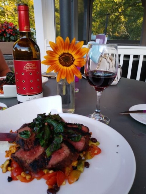 A plate of grilled meat on a bed of vegetables, accompanied by a glass of red wine and a sunflower in a bottle.