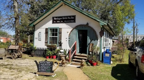 A quaint tea bar with a green roof, wooden door, and outdoor seating, surrounded by trees and a sign for tea offerings.