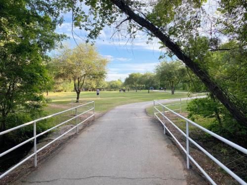 A paved path leads through a park with trees on either side and people in the distance enjoying the open space.