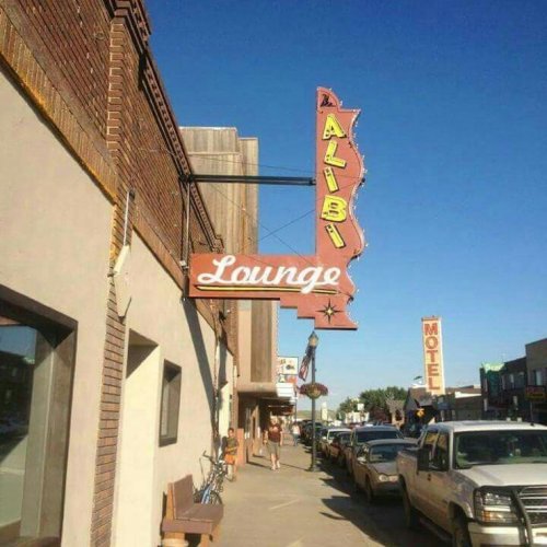 A vintage sign for "Alibi Lounge" hangs above a street lined with parked cars and buildings under a clear blue sky.