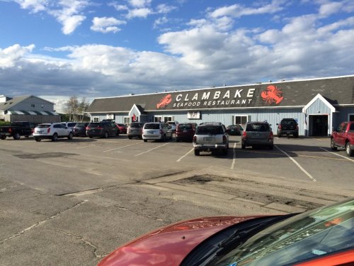Exterior of Clambake Seafood Restaurant with a parking lot filled with cars under a partly cloudy sky.