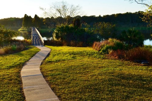 A winding path leads to a wooden bridge over a calm lake, surrounded by lush greenery and trees in the background.