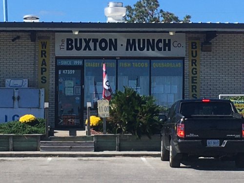 Exterior of The Buxton Munch Co. restaurant, featuring a sign with menu items and a parked black truck in front.