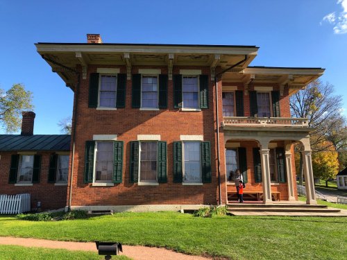 Historic brick house with green shutters, a porch, and a well-maintained lawn under a clear blue sky.