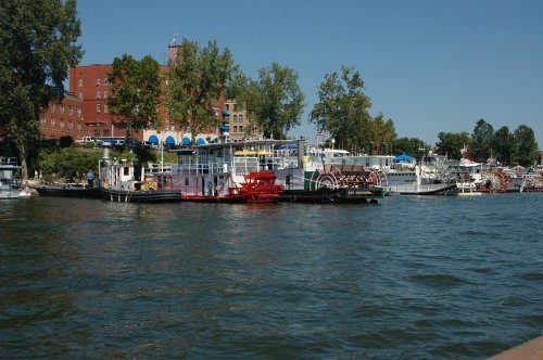 A view of a river with several boats and a waterfront building surrounded by trees under a clear blue sky.