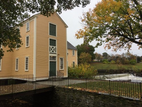A yellow historic building beside a small river, surrounded by trees with autumn leaves on a rainy day.