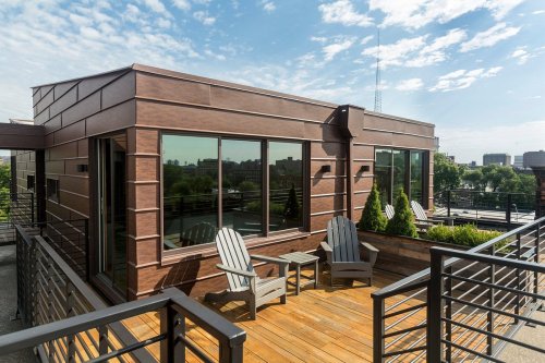 Modern rooftop terrace with wooden deck, two chairs, and greenery, overlooking a city skyline under a blue sky.