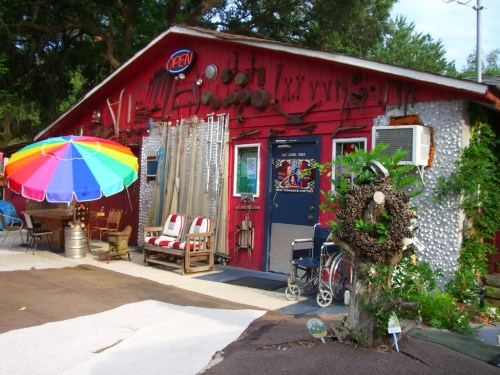 A colorful umbrella shades outdoor seating in front of a rustic red building adorned with tools and decorations.