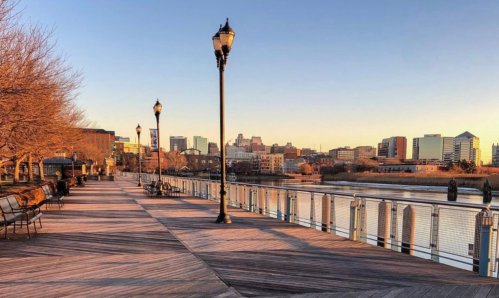 A scenic waterfront walkway lined with benches and street lamps, with city buildings in the background at sunset.