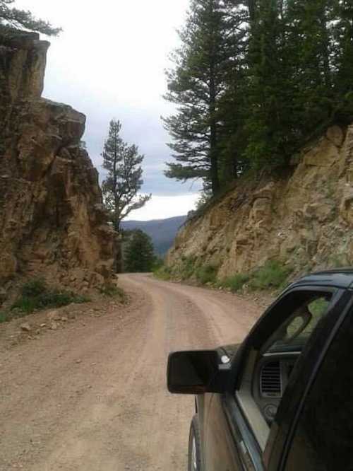 A dirt road winds through rocky terrain and trees, viewed from the open window of a parked vehicle.