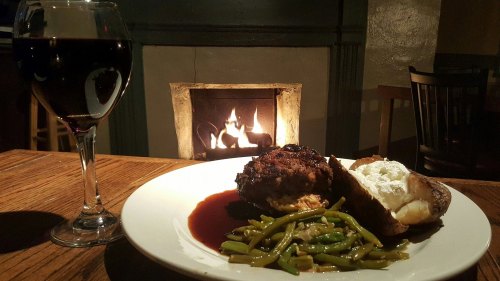 A plate with meat, green beans, and a baked potato, alongside a glass of red wine, with a cozy fireplace in the background.