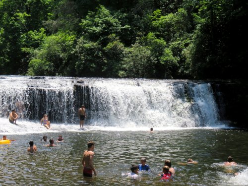 People swimming and playing in a natural pool near a waterfall, surrounded by lush green trees.