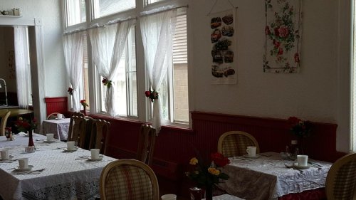 Cozy dining area with tables set for tea, floral decorations, and sunlight streaming through sheer curtains.