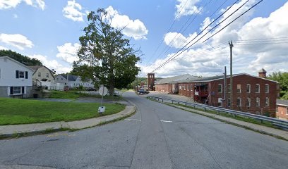 A quiet street scene with houses, a tree, and a brick building in the background under a partly cloudy sky.