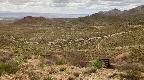 A vast desert landscape with rolling hills, sparse vegetation, and a distant mountain range under a cloudy sky.