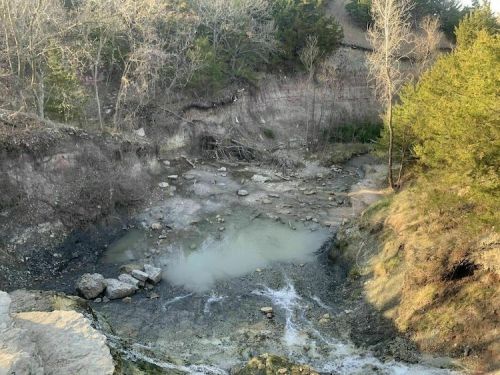 A rocky, dry creek bed surrounded by sparse trees and eroded soil, with a small pool of water visible.