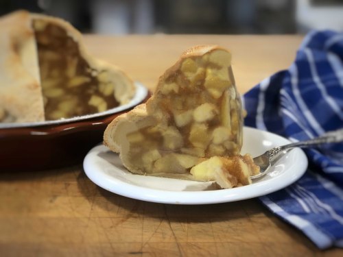 A slice of apple pie on a white plate, with a fork beside it, and a blue checkered cloth in the background.
