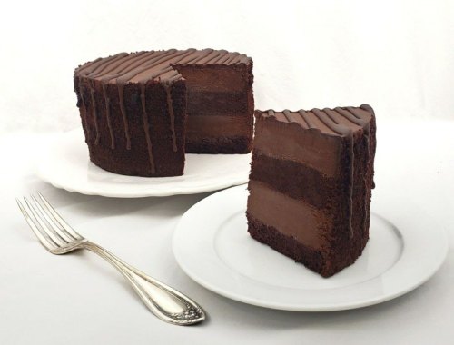 A rich chocolate cake with layers, one slice on a plate beside the whole cake, drizzled with chocolate.