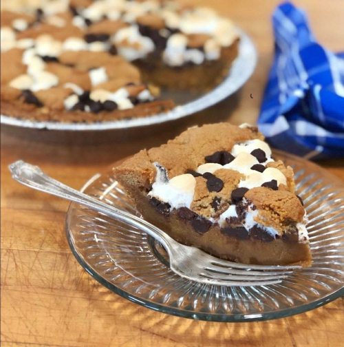 A slice of chocolate chip cookie pie with marshmallows on a glass plate, with the whole pie in the background.
