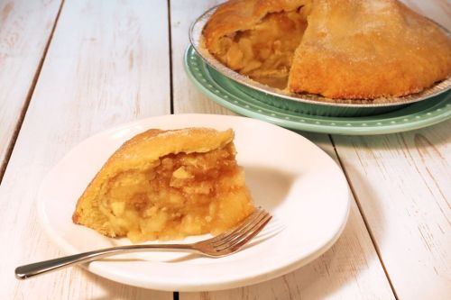A slice of apple pie on a white plate with a fork, next to a whole pie on a green plate.