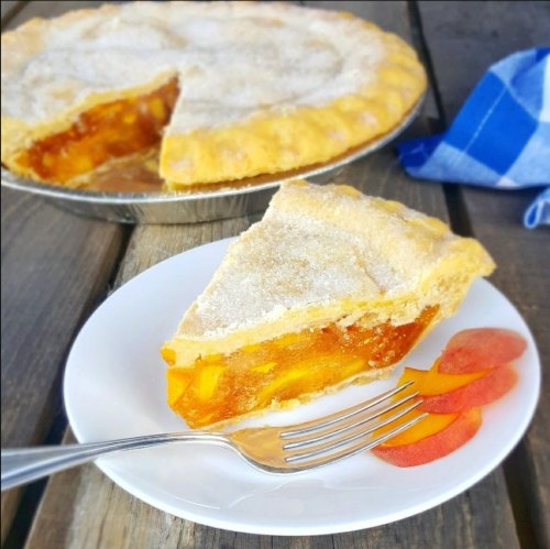 A slice of peach pie on a white plate with a fork, next to fresh peach slices, with the whole pie in the background.