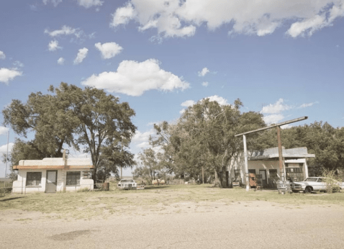 Abandoned buildings and old cars under a blue sky with scattered clouds, surrounded by trees and open land.