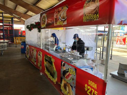 A food stall with colorful banners, featuring two workers preparing dishes and prices displayed for various items.